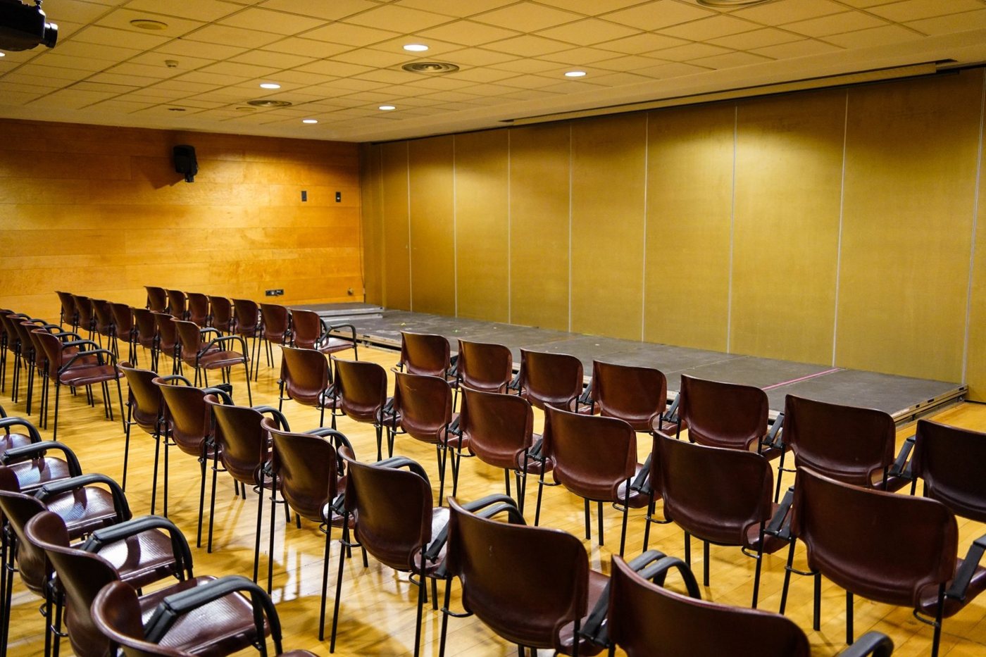 An empty conference room with rows of brown chairs facing a small stage, under an overhead grid-patterned ceiling with recessed lighting. The walls ar